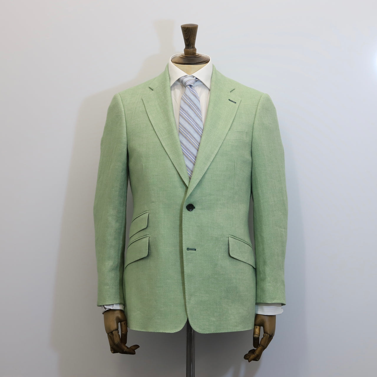 A light sage green linen blazer displayed on a wooden mannequin torso, styled with a white dress shirt and a blue and white striped tie against a plain light gray background.