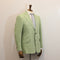 A light sage green linen jacket displayed on a wooden mannequin torso, styled with a white dress shirt and a light blue and white striped tie against a plain off-white wall.