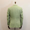 A light green linen-blend suit jacket shown from the back on a wooden dress form mannequin, set against a plain off-white wall.