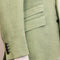 Close-up of a light sage green linen blazer jacket, showing the textured fabric, a flap pocket, and four green buttons on the cuff.