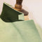 Close-up of a light sage green herringbone fabric garment or textile draped on a dress form, with a darker green collar section and a wooden mannequin neck visible against a soft light background.